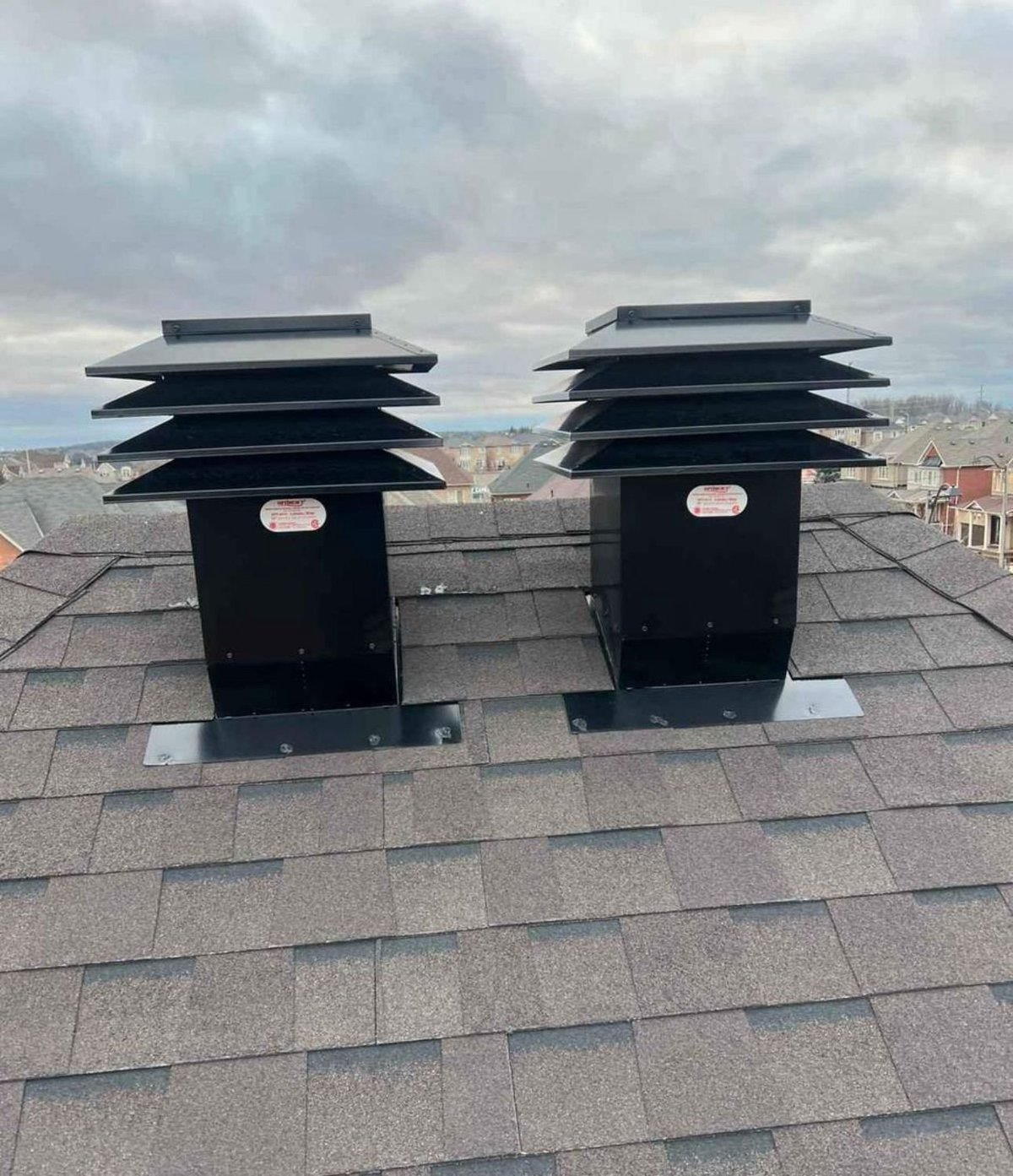 Two black roof ventilation fans mounted on asphalt shingles with residential buildings visible in the background