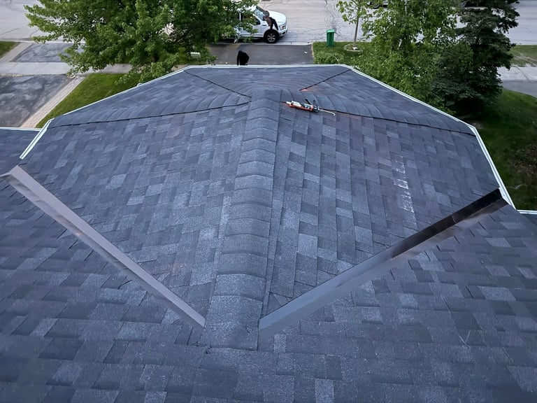 Aerial view of a house roof with gray asphalt shingles, showing multiple roof sections and surrounding trees and driveway