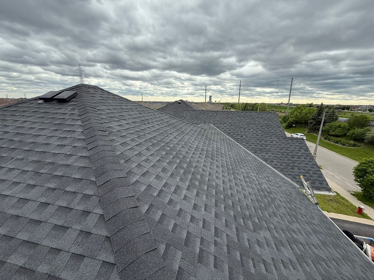 Aerial view of a dark gray asphalt shingle roof with multiple peaks overlooking a residential neighborhood under a cloudy sky