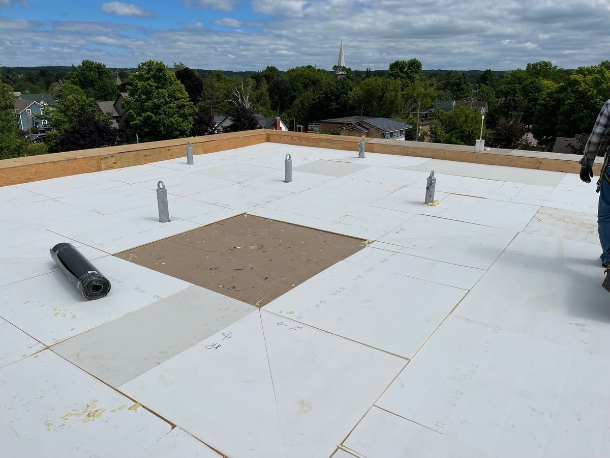 Flat roof with white membrane installation, metal vents, and rolled roofing material visible against a suburban landscape